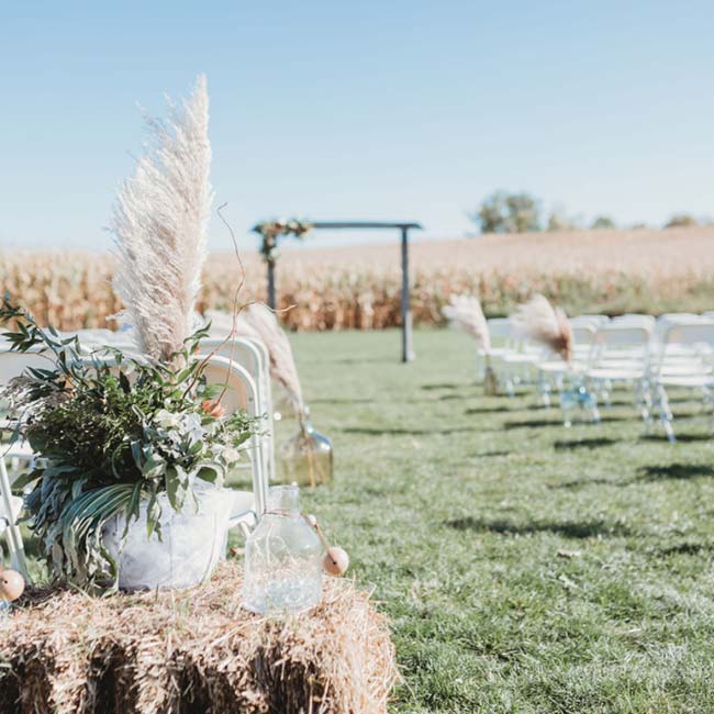 Outdoor barn wedding setup with white chairs, hay bales, greenery, and a wooden arch in a field.