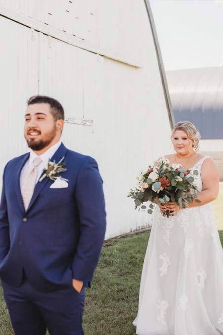 Bride with a bouquet walks toward groom in blue suit for a Barn Wedding first look outside.