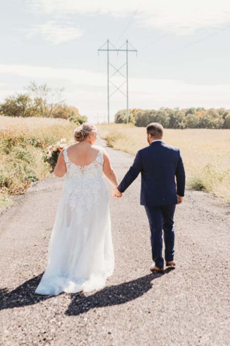 A bride and groom walk hand-in-hand down a sunny Minnesota road near their barn wedding venue.