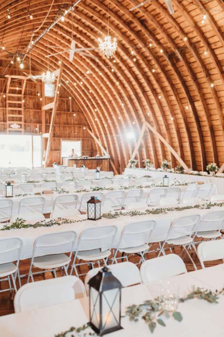 Rustic barn wedding venue with white tables, chairs, greenery, lanterns, and string lights.
