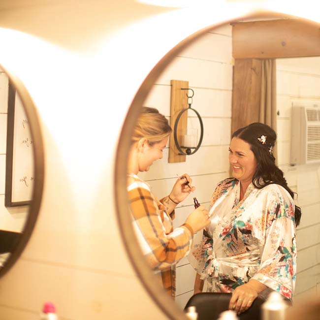 Two women smile in a mirror as one helps the other get ready for a rustique barn wedding ceremony.