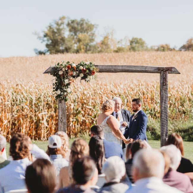A couple weds outdoors under a floral arch at Rustique Barn, with guests in a Minnesota field.