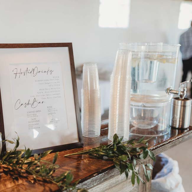 Framed drink menu, stacked cups, water dispenser, and greenery at a Rustique Barn wedding.
